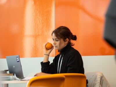 Eine junge Frau sitzt vor einem Laptop und hält eine Orange in ihrer Hand. Hinter ihr ist eine orange Wand.